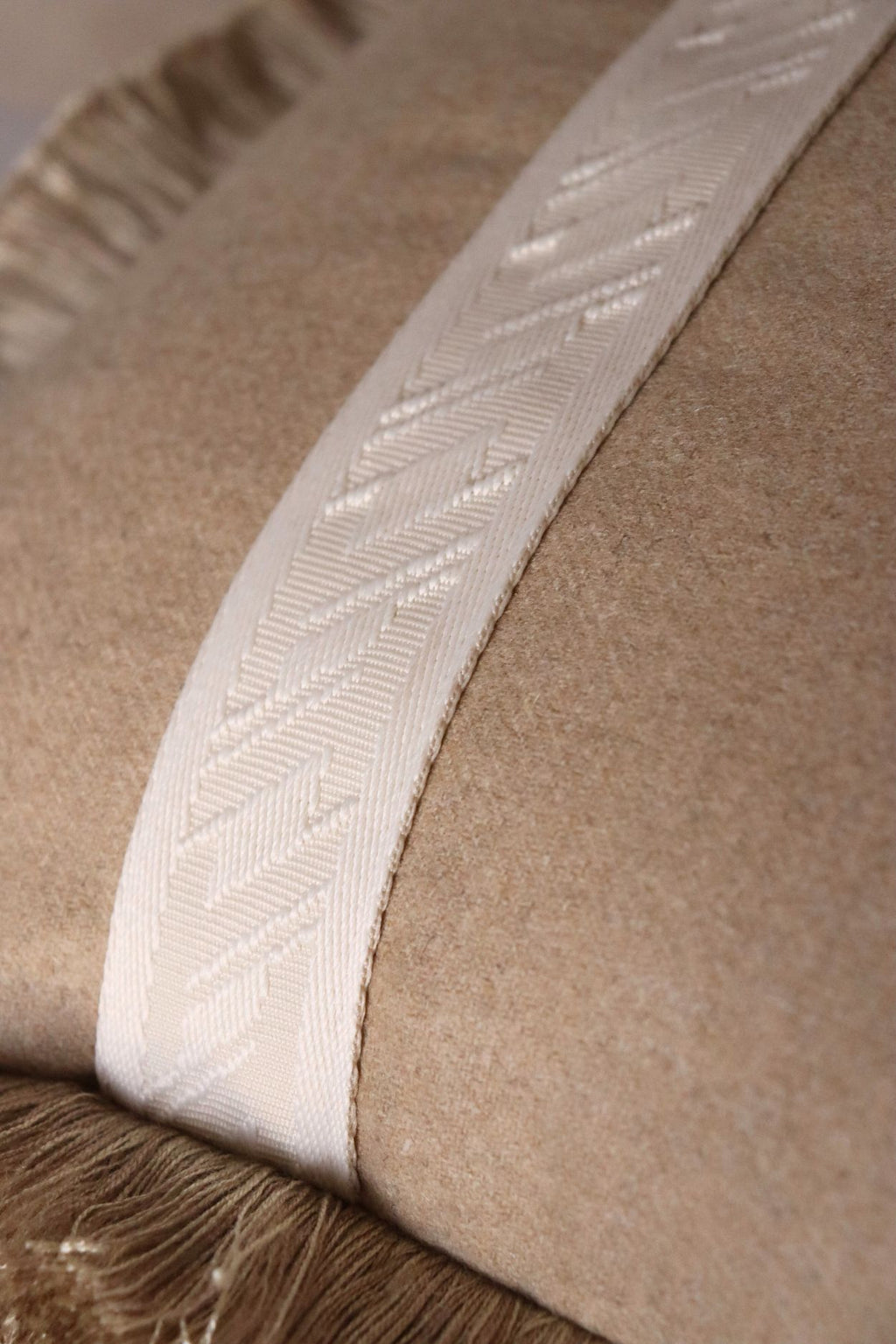 De stof van een wollen beige sierkussen met witte band en beige franjes.