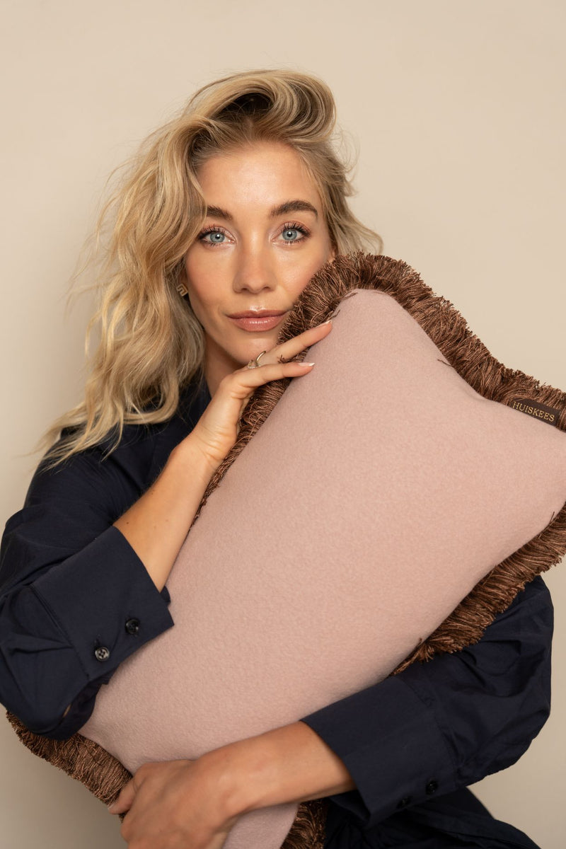 Een blonde vrouw houdt een roze sierkussen met lichtbruine franjes vast.