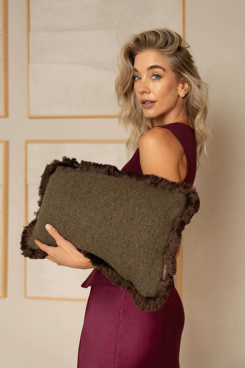 Een blonde vrouw houdt een donkergroen sierkussen met bruine franjes vast.