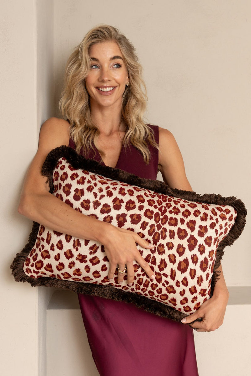 Een blonde vrouw houdt een rechthoekig sierkussen met luipaardmotief en bruine franjes vast.