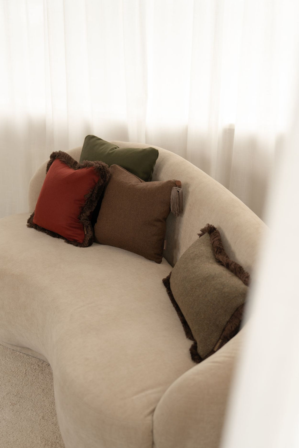 Twee groene, een rood en een bruin sierkussen met kwastje liggen op een beige bank.