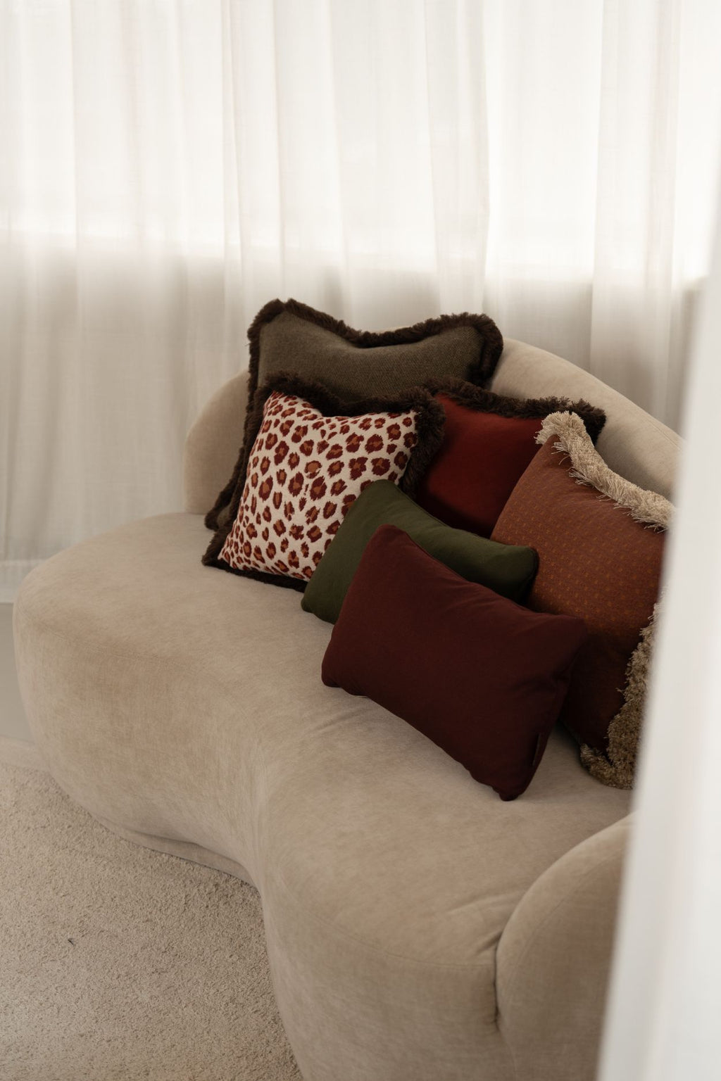 Groen sierkussen met franjes, rode sierkussens met print en franjes en een oranje sierkussen liggen op een beige bank.