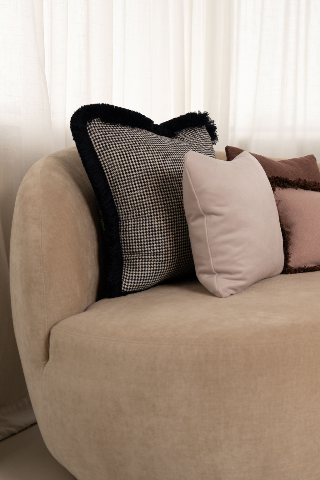 Een donkerblauw sierkussen met pied-de-poule motief en blauwe franjes ligt met een lichtroze sierkussen op een beige bank.