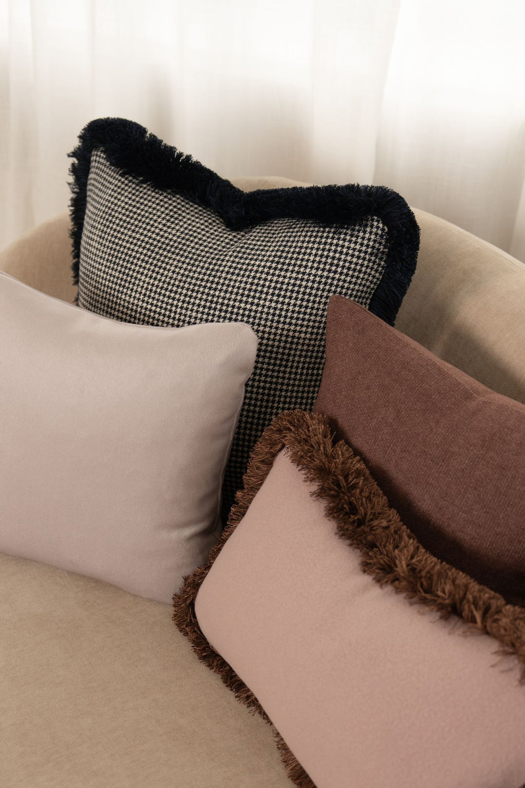 Een donkerbkauw sierkussen met franjes ligt op een beige bank met roze sierkussens met en zonder franjes.