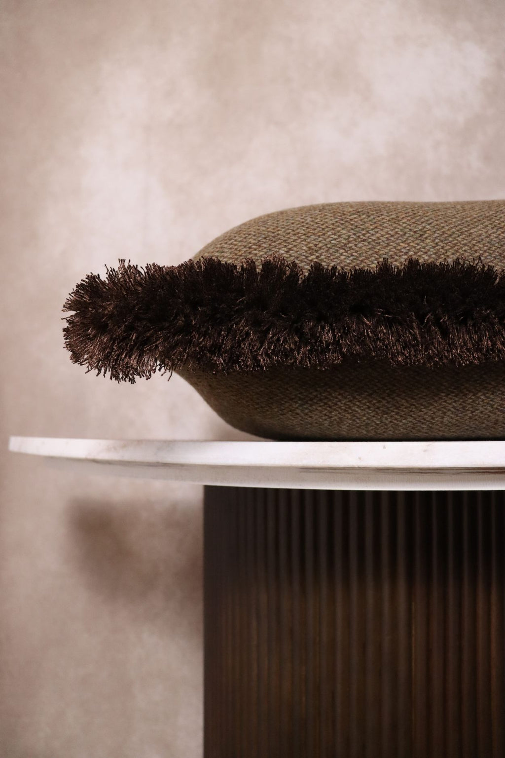 Een donkergroen sierkussen met bruine franjes ligt op een tafeltje.