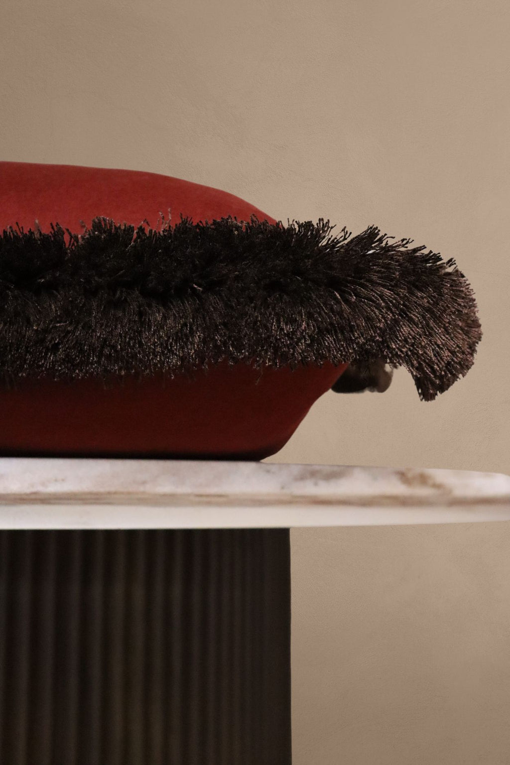 Een rood sierkussen met bruine franjes ligt op een tafeltje.