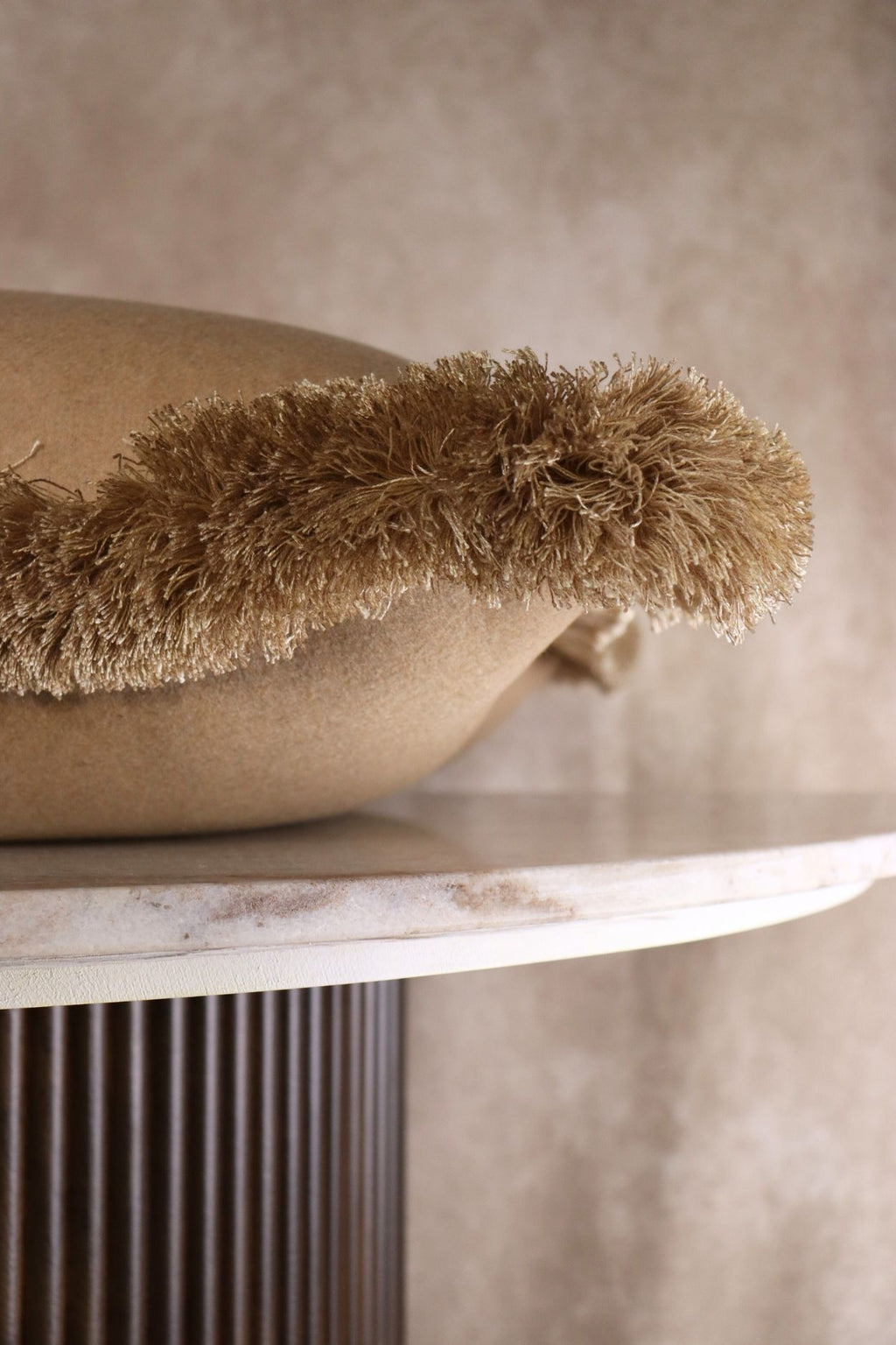 Een beige sierkussen ligt op een tafeltje zodat de beige franjes goed zichtbaar zijn.