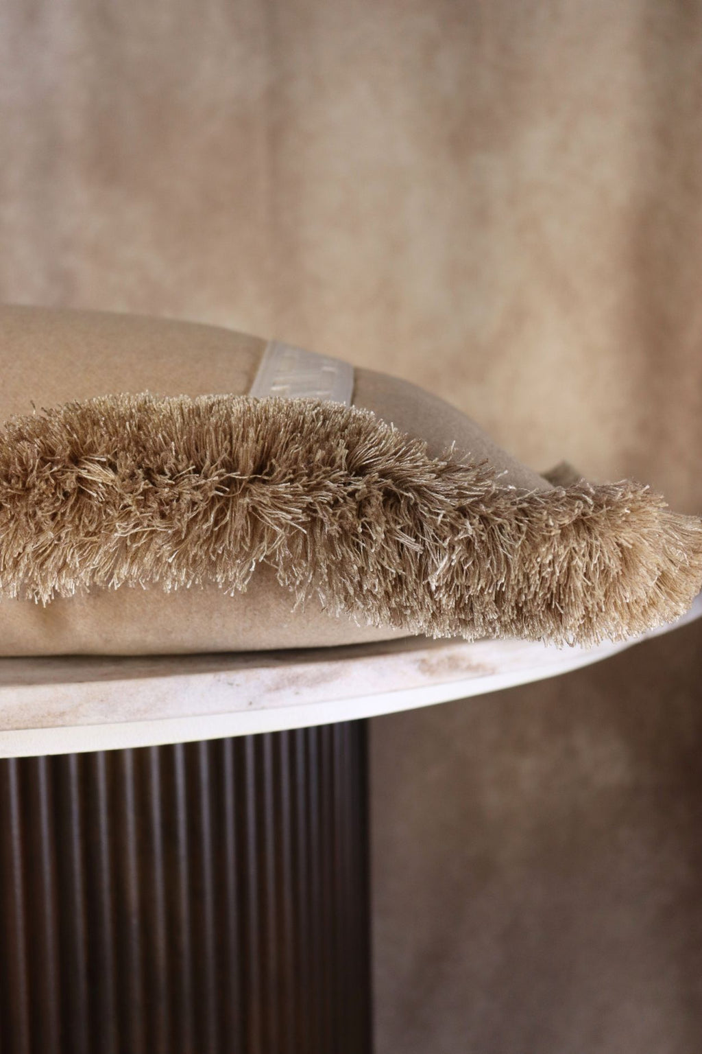 Een beige sierkussen met witte band en beige franjes ligt op een tafeltje.