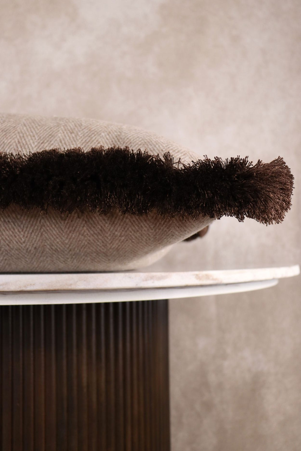 De bruine franjes van het bruin-beige sierkussen met visgraatmotief is goed zichtbaar omdat het kussen op een tafeltje ligt.