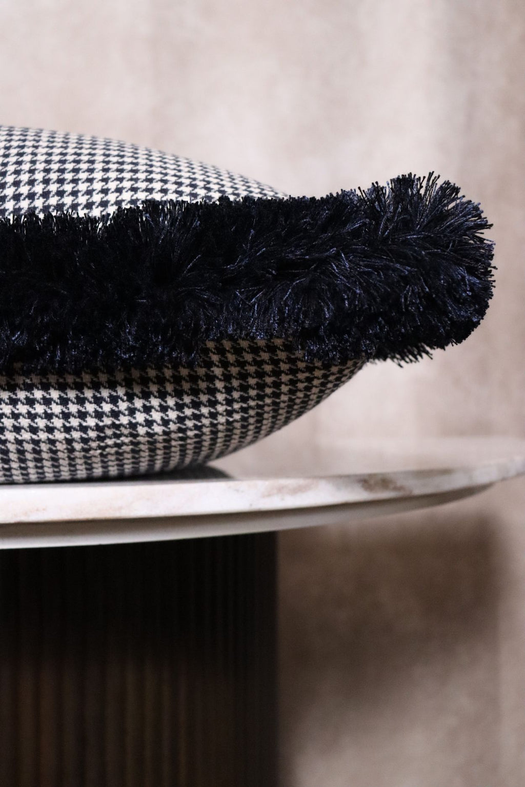 Een donkerblauw sierkussen met franjes en pied-de-poule motief ligt op een tafeltje zodat de franjes goed zichtbaar zijn.