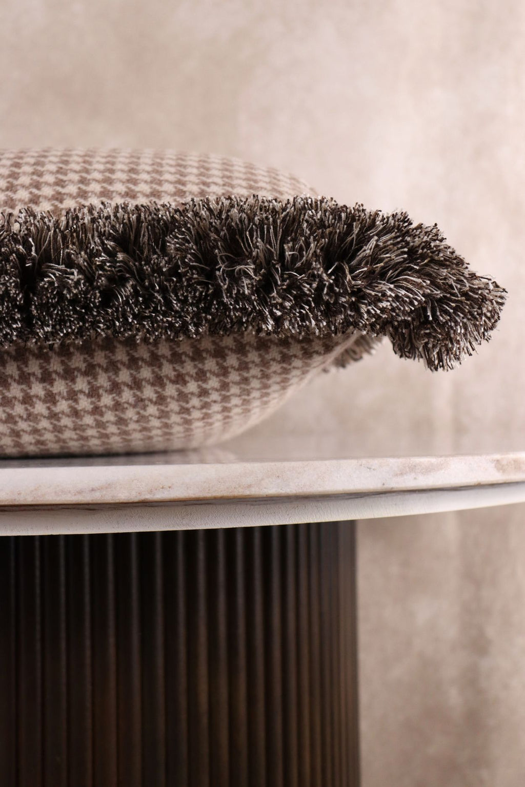 Een lichtbruin sierkussen met pied-de-poule motief ligt op een tafeltje zodat de franjes goed zichtbaar zijn.