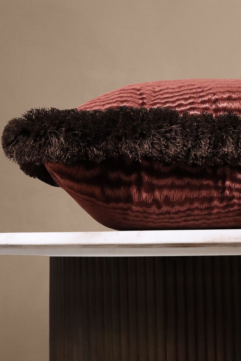 Een luxe donkerroze sierkussen met bruine franjes ligt op een tafeltje.