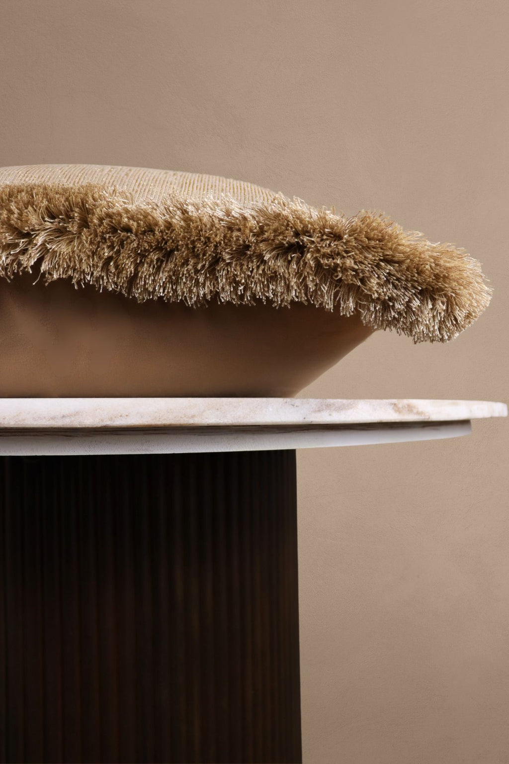 Een beige sierkussen met beige franjes ligt op een tafel.