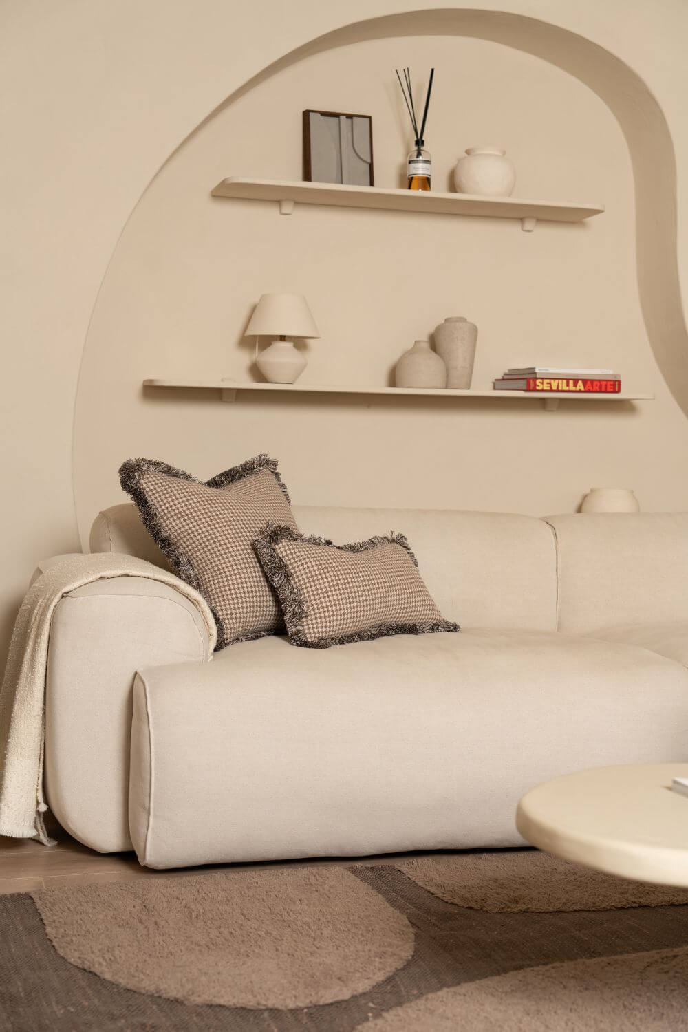 Twee beige sierkussens met bruin franjeband liggen op een witte bank.