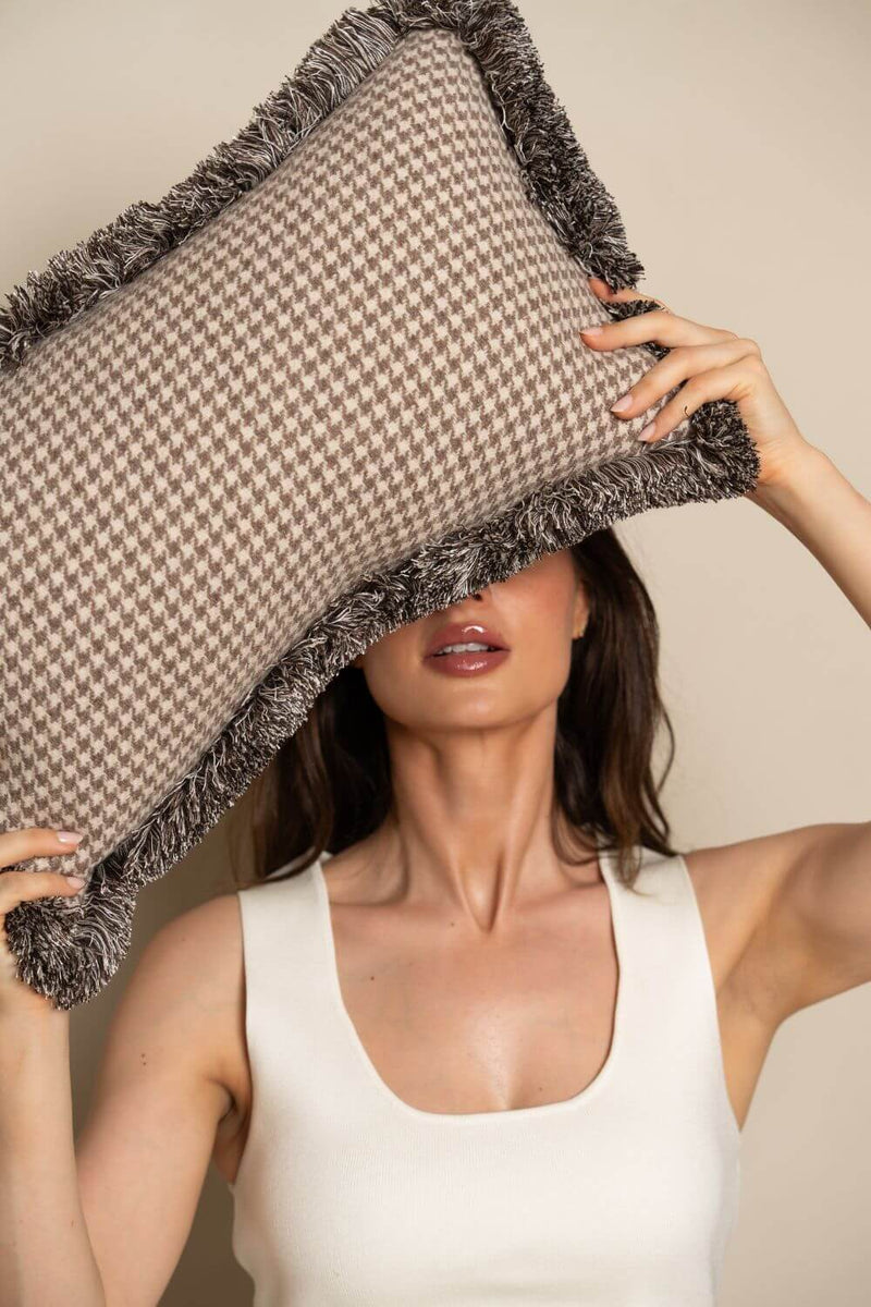 Een vrouw houdt een beige sierkussen met pied-de-poule motief voor haar hoofd. Het sierkussen heeft een tweekleurig franjeband.