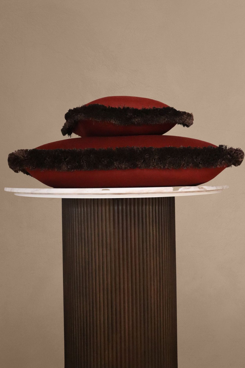 Twee rode sierkussens met bruine franjes in verschillende formaten liggen op elkaar op een tafeltje.