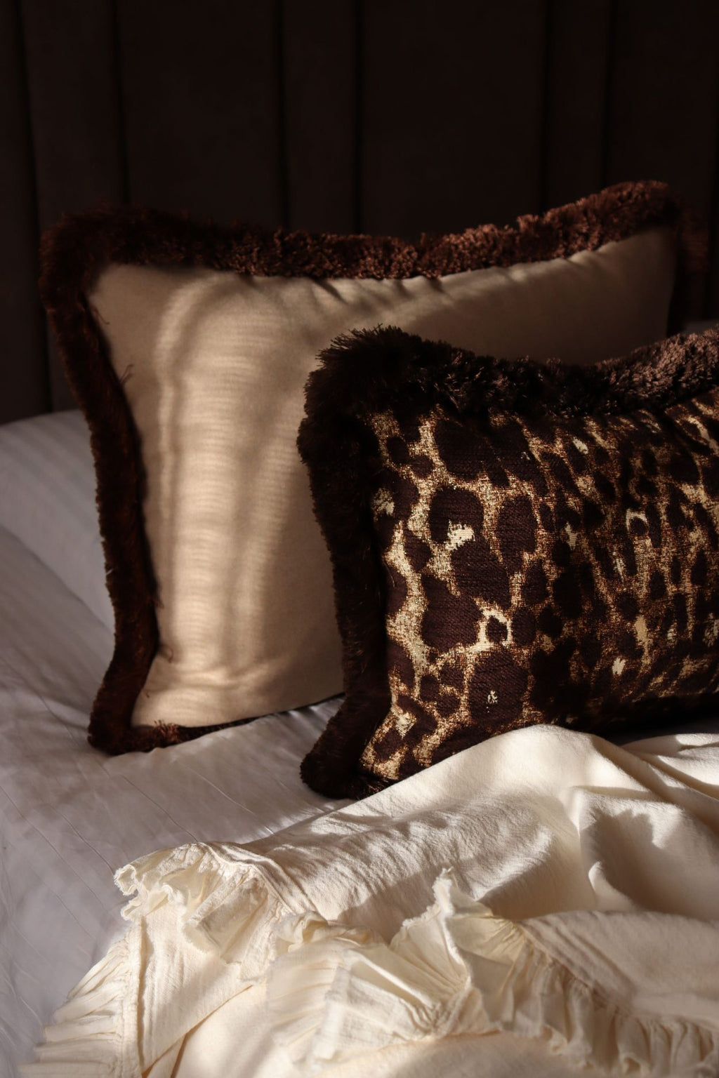 Een groot beige sierkussen met bruine franjes en een bruin sierkussen met luipaardmotief liggen op bed.
