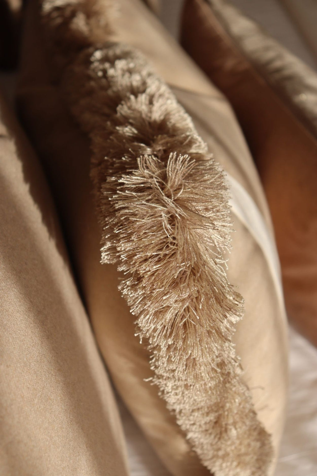 De beige franjes van een rechthoekig sierkussen op een bed.